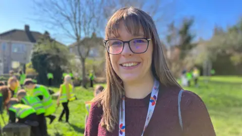 A young woman with light brown hair and glasses is smiling at camera in a park with children in hi-ves vests behind her. 