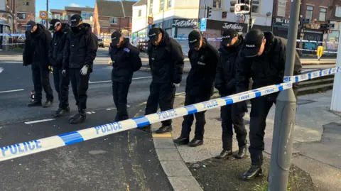 Tom Ingall/BBC A row of police officers with their heads down are all looking at the floor for forensic evidence. They are surrounded by police tape. 