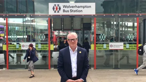 LDRS The West Midlands mayor Richard Parker, a middle-aged man with white hair and spectacles, stands in front of a set of glass doors under a sign reading "Wolverhampton Bus Station". He is wearing a blue suit jacket and a white open-necked shirt.