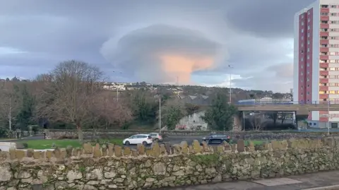 Rab Jones A stone wall, with the view of a town in the distance and a high rise building to the right. The sky has a large cone shaped cloud.