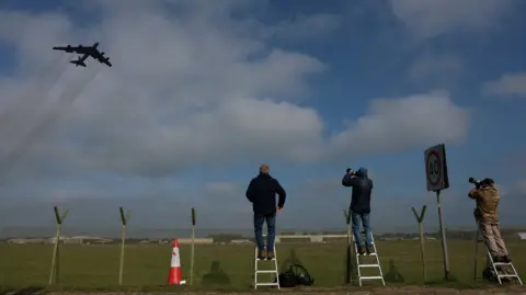 Reuters Three plane-spotters on short step ladders stand at the perimeter fence of an airfield with a B-52 plane flying overhead in a blue sky with white clouds.
