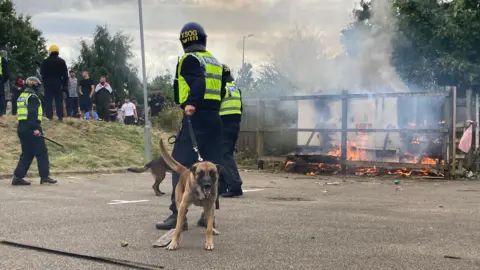 BBC/Oli Constable Three police officers and two police dogs can be seen in front of a fire. A group of men is standing on a grass verge next to it.