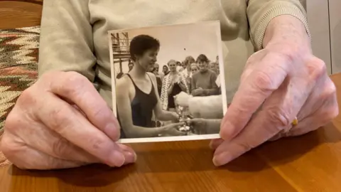 A pair of hands, holding a black and white picture of a lady with short hair and a black bathing suit receiving a trophy in front of a crowd.