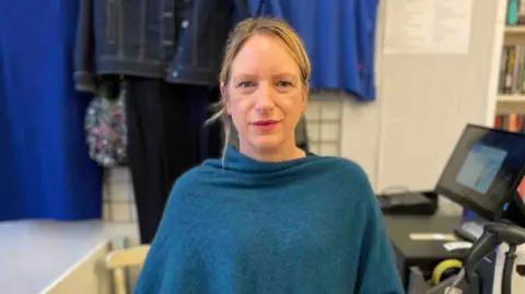Blythe House Hospice Senior Retail Manager Marie Brown is pictured wearing a blue shawl. She has blond hair which is tied back. Behind her is a till