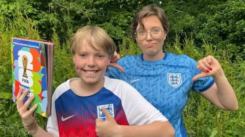 Jack, an 11-year-old boy with blonde hair, holds a World Cup 2026 Panini sticker book, a brightly coloured logo with the tournament trophy on it. He is wearing a white, blue and red vintage England shirt. He has his thumbs up and is smiling. Sophie, a woman with short dark hair and wearing glasses stands behind him. She is wearing a blue patterned women's England shirt. Her face is perplexed and she has her thumbs down. 