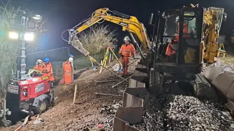 Network Rail At night, a mechanical digger rests on stones on an embankment which is supported by sheet metal piles. Uniformed workers and other machinery surround it, illuminated by floodlights.