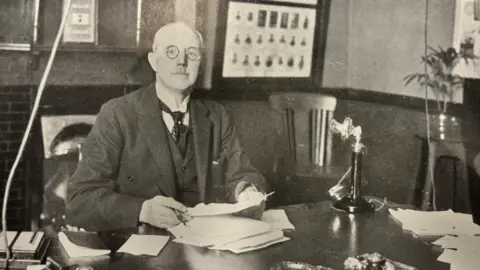 Hull History Centre An old photo, in black and white. It shows a man looking at the camera. He's wearing glasses and a suit. He's sitting at a table, and is holding numerous papers and a pen. He's sat in an office. 