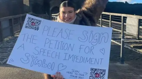 Jessie Yendle Jessie Yendle holding white placard with QR codes and message saying please sign the petition for a speech impediment symbol