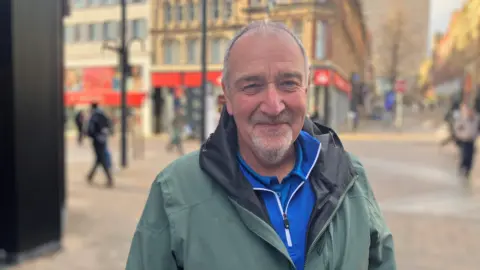 A man with short grey hair and stubble is wearing a green coat over a blue top