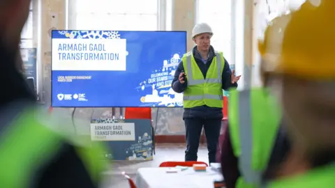 Armagh, Banbridge and Craigavon Council Kieran Carlin addressing members of the public inside Armagh Gaol during a public engagement session. He is wearing a white hard hat and a yellow high-viz vest over dark trousers and dark jacket. He is gesturing with both hands as he talks. A large TV screen behind him shows a sign which reads "Armagh Gaol Transformation" on a bright blue background.