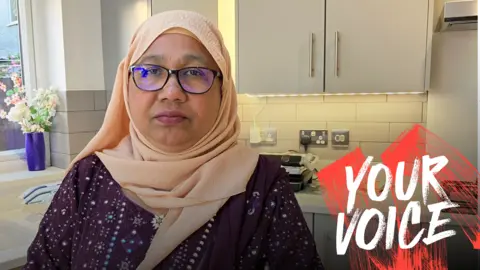 BBC A woman in a peach hijab and glasses stands in a modern grey-hued kitchen. 