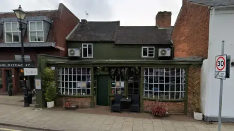 Google The terraced bar is painted olive green with large windows either side of its entrance and two seats outside. There are some floral/greenery decorations at the front as well.
