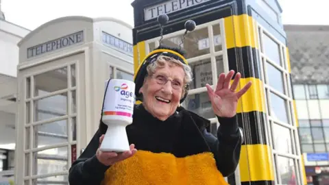 Age UK Jean Bishop wearing glasses and dressed in her bee outfit stands in front of a telephone box painted black and yellow. She is holding a white Age UK collecting tin