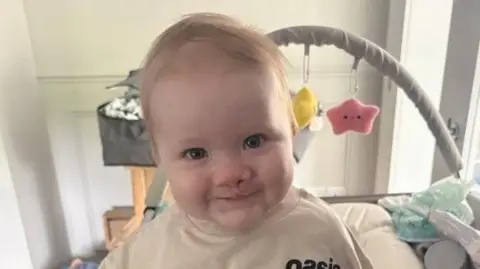 BBC A baby, Eli. He is wearing a beige t-shirt. He has a grey baby activity arch behind him with yellow and pink stars. 