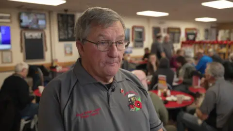 Denis wears a grey top with a Poppy Appeal logo on it. Other veterans are seated at tables behind him.