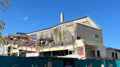 A former cinema building, with a cream-coloured exterior, has been partially demolished revealing the inside of the building with lots of debris lying on one of its floors, with a blue sky behind it