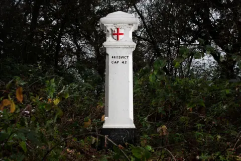 Historic England A white post with a pointed top with an England flag on the front surrounded by trees