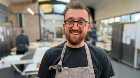 A young bearded man wearing a dark T‑shirt and a grey apron with the “Six Chimneys” logo stands inside a bright, industrial bakery workspace, with stainless‑steel equipment and preparation tables visible in the background.