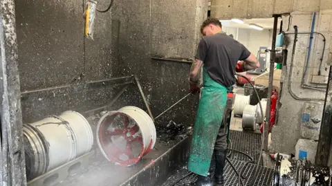 JOZEF HALL/BBC Keiran Thomas strips old alloy wheels. He is photographed from the side and he is wearing a green apron and black T-shirt and is working in a garage. 