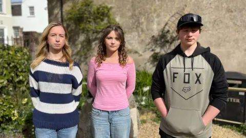 Left - a girl with shoulder length blonde hair, wearing a jumper with blue and white horizontal stripes. Centre - a girl with shoulder length brown hair, wearing a pink top. Right - a boy with short dark hair, wearing a black baseball cap and a hooded jumper.