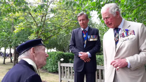 Chris Redston Bill Redston wearing a navy jacket and hat looking up at King Charles wearing a pale suit and medals, with another man in a suit and medals standing with his hands clasped in the background