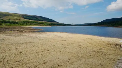 PA Media The reservoir has shrunk exposing a sandy coloured dry bed. Green hills can be seen rising behind