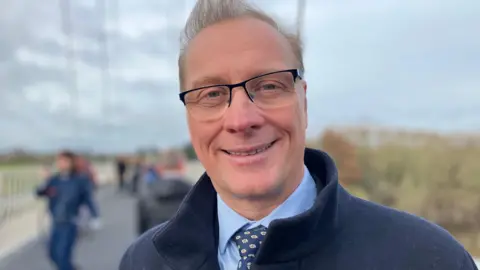 Worcestershire County Councillor Marc Bayliss, a man with short grey hair and glasses, smiles at the camera in a shot taken outside. He is wearing a light blue shirt and a dark blue tie under a dark blue coat.