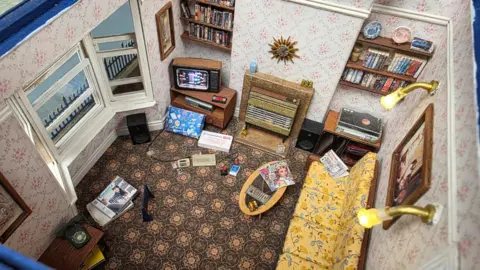 Lee Smithson A detailed miniature model of a living room designed in a retro style. The room has patterned wallpaper with small floral prints and a carpet featuring an intricate geometric design. Against one wall, there is a yellow sofa with a floral pattern, and in front of it is a small oval wooden coffee table with magazines placed on top.
On the opposite wall, there is a fireplace with a decorative sunburst clock above it, flanked by shelves filled with books and DVDs. A small television sits on a wooden stand near the fireplace, and there are speakers positioned on either side. Various items, including boxes, newspapers, and a remote control, are scattered on the floor.
The room also features framed pictures on the walls, a window with a scenic painting visible outside, and two wall-mounted lights providing illumination.