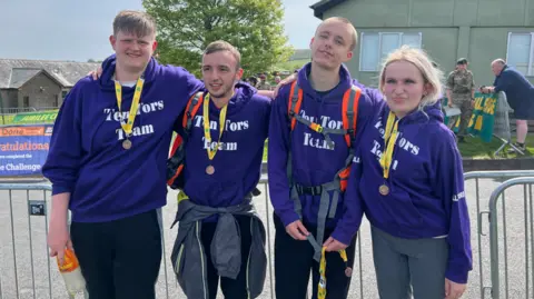 A group of four people stand in a row, side by side. They are all wearing purple hoodies with 'Ten Tors Team' written on the front. They each have a medal. 