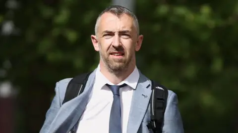 Liam McBurney/PA Wire Daithí McKay walking towards a camera on a street during an earlier hearing in the case.  He has short, greying hair and a beard. He is wearing a light grey suit, a pale or white shirt and a black tie.  He is carrying a black backpack with a strap over each shoulder. 
