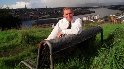 Lions Rugby Ken Goodall, sits on a black park benc, in a grassy field. Behind him is a views of the city of Derry, including the river Foyle.
