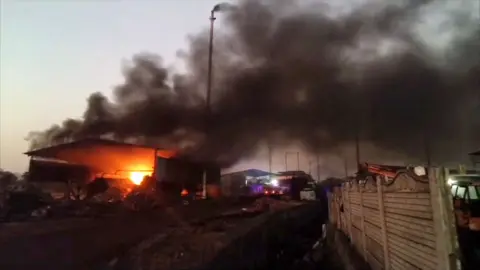 A still from footage of an explosion in a pyrolysis plant. Flames can be seen inside an open building a thick black plumes of smoke fill the sky above. 