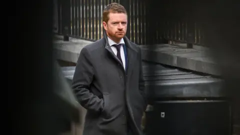 Morgan McSweeney, the prime minister's former chief of staff, walking down Downing Street. He has a serious expression and wears a dark grey coat with the collar turned up over a white shirt with a black tie. He has short hair and a very short beard.