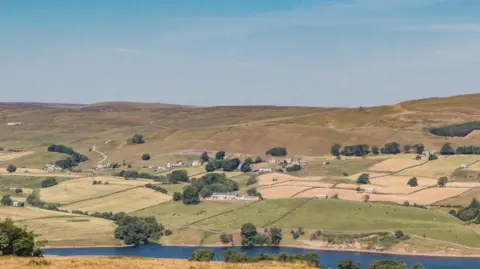 A landscape of rolling hills, with fields divided by stone walls leading to more open land. To the foreground runs a river, with trees fringing the banks in parts. Scattered around are stone houses.