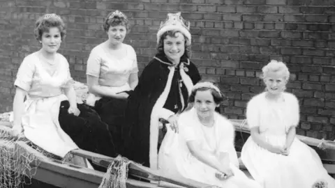 Berwick Town Council Three women and two young girls sit in a boat together. Netting is visible on either side of the boat which is moored on dry land next to a wall. All five are wearing white dresses ,with the woman in the centre wearing a cape and a crown 