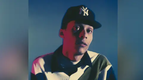 Man with a baseball cap and collared long sleeve shirt, with a blue background