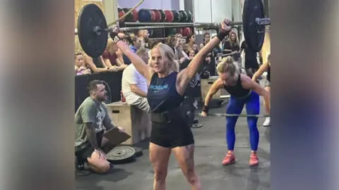 NHS Humber Health Partnership Amy Carberry inside a gym, holding a barbell, with a weight on either side, above her head. She is wearing a navy vest with blue Nike branding reading "ROGUE" in capital letters and black shorts. She has long blonde hair tied back. Other people are behind her lifting weights and some people are spectating. 