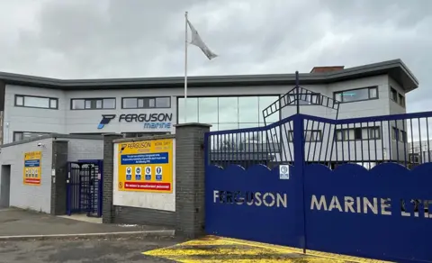 Gerbang galangan kapal biru dengan Ferguson Marine memotong ke dalam desain. Di latar belakang adalah gedung kantor abu -abu dengan logo yang sama. 