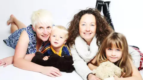 Kerry and Julie with their young children, Ben and Olivia in a professional family photograph pictured lying on their fronts on the floor with a white background. 