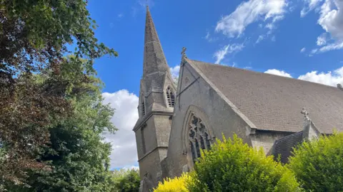 A view looking upwards at a stone church and its church spire. There are lime green bushes in the foreground and above is a blue sky with white clouds.
