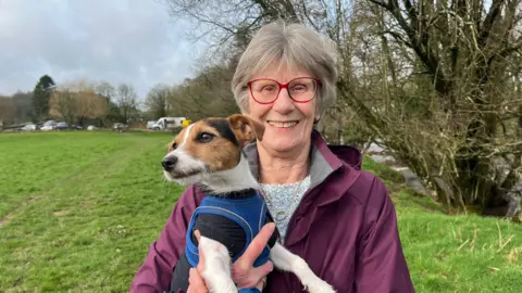 A woman stands in a field smiling at the camera, holding a Jack Russell dog. She's wearing a purple rain coat and red glasses. The dog has a blue harness on. On the right is a is flowing river.