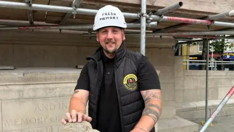 The photo shows a man smiling at the camera. He's wearing a black gilet, back polo shirt and white hard hat. He's standing in front of a warm memorial, and has his hand on a piece of stone. The memorial is covered in scaffolding. 