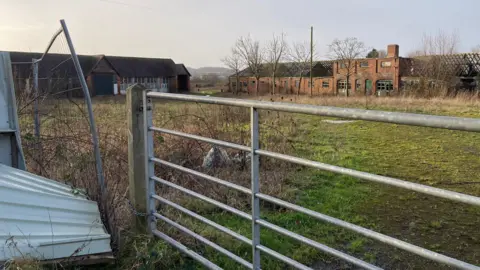 A rather unkempt field with two buildings in the background. The field is closed off by a gate and there is an overgrown patch of brambles.