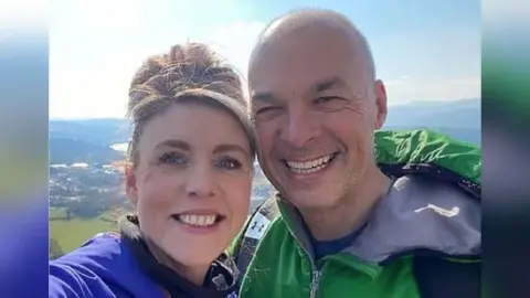 Rebecca Hutt (left) and Det Supt Steven Williams (right) are photographed together from the shoulders up. They are both smiling and looking at the camera. They are outdoors on a sunny day at the top of a high view point. 