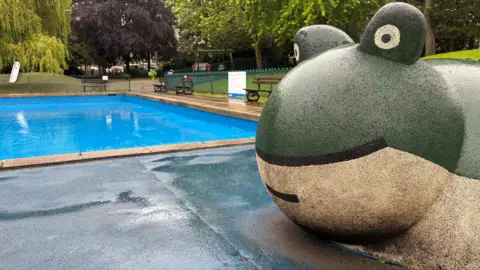 A paddling pool in a public park - a large rectangle of water, surrounded by green metal fencing. In the foreground, a large frog sculpture can be seen, and swings and other play equipment can be seen in the background.