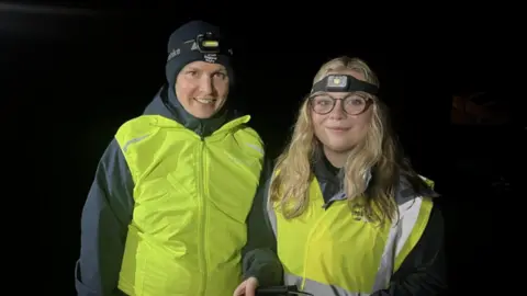 A woman with a black hat and black coat alongside a woman with long blonde hair and glasses. Both are wearing yellow reflective jackets