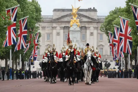 LPhot Mathew Bradley / MOD The procession of military personnel riding horses parade down the Mall with union jacks and green foliage lining the street and Buckingham Palace in the background.