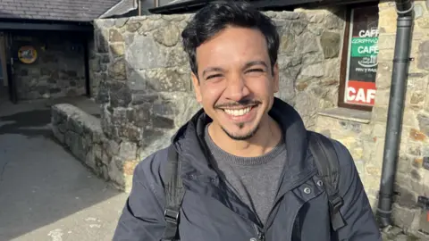 Mohiuddin Kamal, a man with a moustache, chin beard and short brown hair smiling in front of a stone building