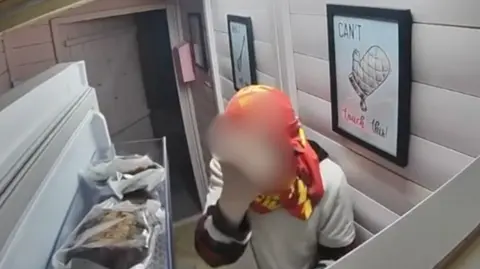 An image from CCTV shows what appears to be a young man with a red and yellow football shirt wrapped around his head like a balaclava. He has opened up a fridge with cakes in wrappings in the foreground. The person's hand has been blurred out, as he makes a rude gesture at the cctv.