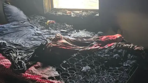 Cambridgeshire Fire and Rescue Service A bedroom with charred walls and a window in the centre. The covers and mattress on the bed have been burned, exposing the metal springs underneath. 